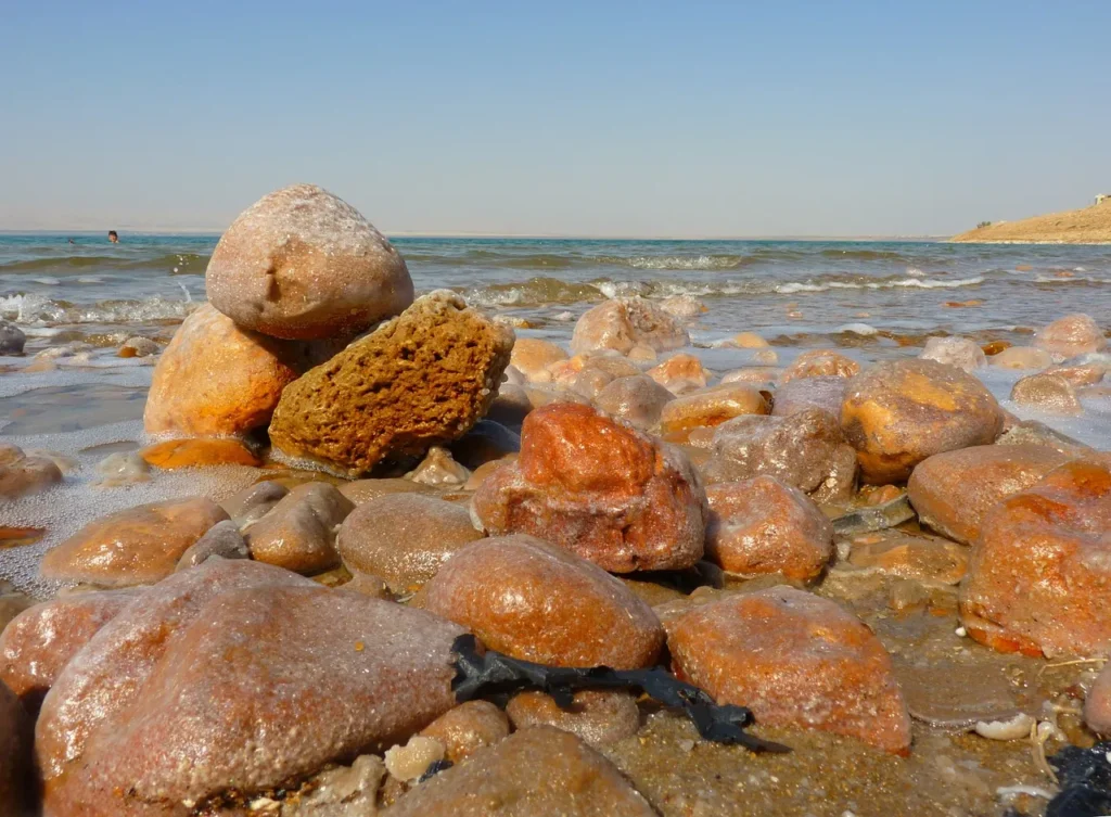 Salt-encrusted rocks and crystal formations on the shore of the Dead Sea with clear blue water and distant hills under a bright sky.