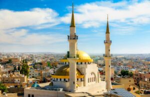 A stunning aerial view of a large mosque in Madaba, Jordan, featuring prominent golden domes and two tall, slender minarets under a clear blue sky with scattered clouds, overlooking a densely populated urban landscape.