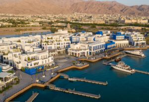 Aerial view of Ayla Oasis Marina Village in Aqaba, Jordan, featuring white and blue buildings, a marina with yachts, and mountains in the background.