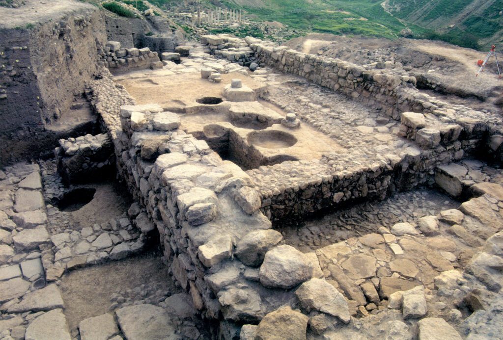 n aerial view of archaeological excavations at the ancient city of Pella in Jordan, showing stone foundations, walls, and excavated pits of a historical structure, possibly a temple or domestic area, with a hilly landscape in the background.