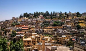Panoramic view of Al-Salt, Jordan, a city built on hillsides with densely packed historic stone buildings and a prominent minaret, under a clear blue sky.