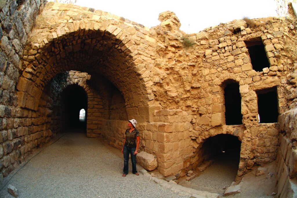 A woman stands within the stone-vaulted halls and arched passageways of Kerak Castle, a historic Crusader fortress in Jordan, with sunlight illuminating the entrance of a dark tunnel.
