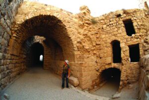 A woman stands within the stone-vaulted halls and arched passageways of Kerak Castle, a historic Crusader fortress in Jordan, with sunlight illuminating the entrance of a dark tunnel.