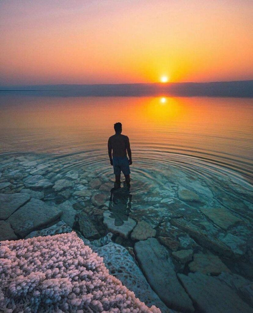 Man standing in the Dead Sea during a vibrant sunset, with salt crystal formations visible in the foreground