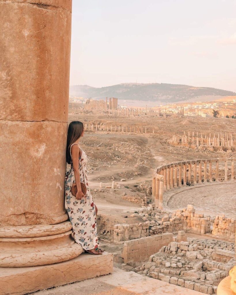 A woman in a floral dress stands by a large stone column, looking out over the ancient ruins of the Amman Citadel with its scattered columns and structures under a clear sky