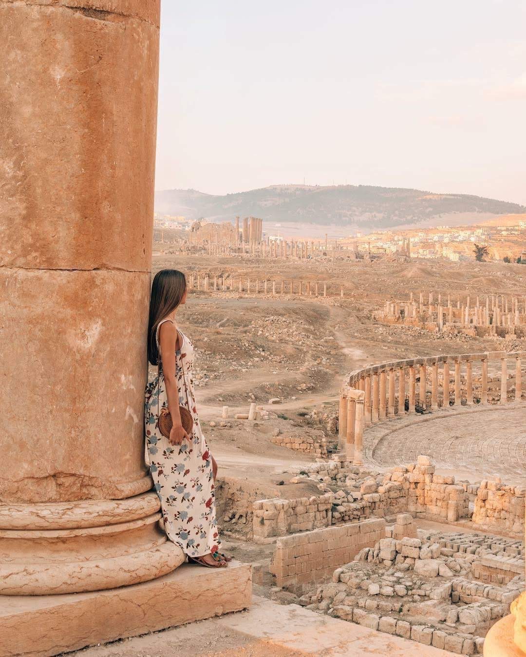 A woman in a floral dress stands by a large stone column, looking out over the ancient ruins of the Amman Citadel with its scattered columns and structures under a clear sky